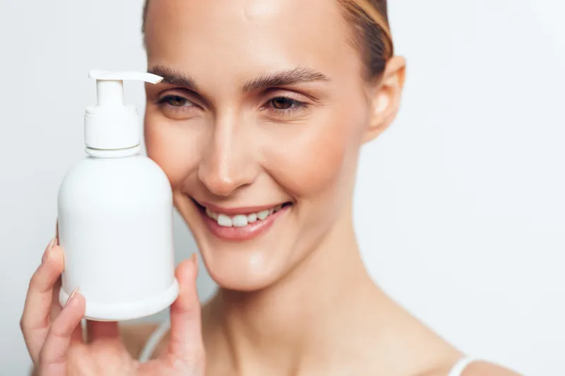 Smiling woman holding a white plastic pump bottle close to her face on a white background. Concept of skincare, beauty, hygiene, and personal care with soft natural lighting.