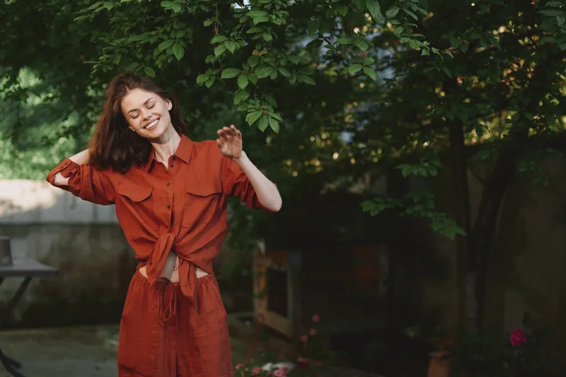 A joyful woman outdoors in a rust dress, smiling, in a sunlit garden. A candid lifestyle portrait showcasing casual fashion, greenery, warmth, and relaxed confidence