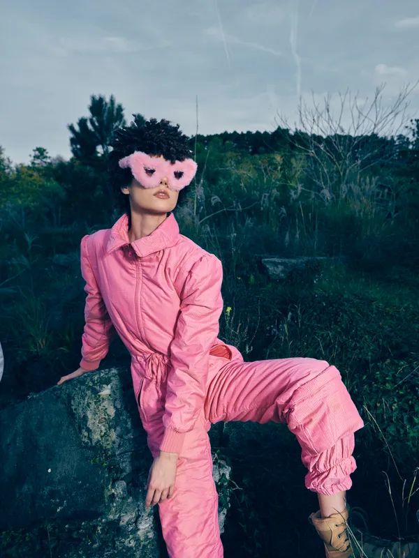 Woman in a pink jumpsuit and masquerade mask posing on a rocky ledge outdoors, bold fashion portrait with dramatic color and confident stance amid natural scenery during daylight