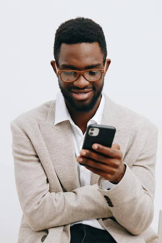 Man using smartphone, smiling, casual beige blazer, glasses, African descent, modern lifestyle, tech, communication, urban, warm clothing, indoors, positive mood, portrait. People lifestyle concept.
