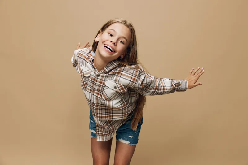 Girl in plaid shirt and denim shorts smiling happily with arms outstretched standing against beige background, casual youth fashion and joy expression.