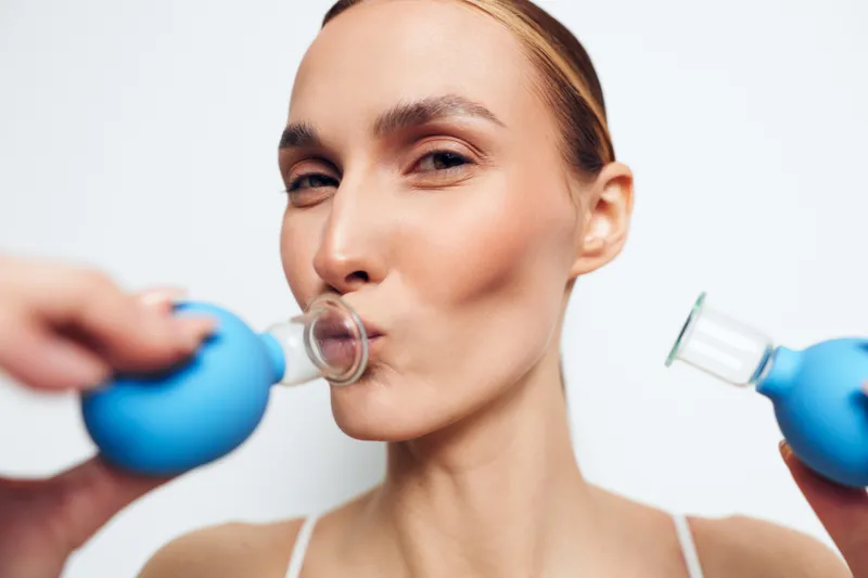 Young woman using facial cupping therapy with blue silicone cups on her cheeks. Close-up on white background. Concept of skincare, beauty treatment, and natural wellness.