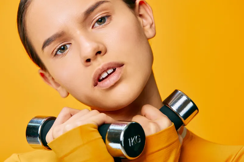 Close-up of a woman with dumbbells against a bright yellow backdrop, showcasing focused fitness and athletic training for strength, balance, and health in a modern sport vibe.