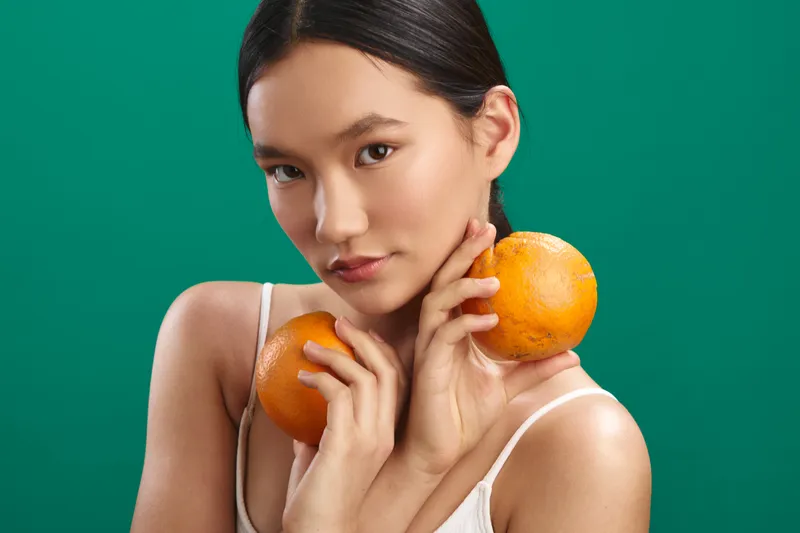 asian woman holds oranges against a vibrant green backdrop, creating a fresh, playful mood. she gazes softly at the camera, balancing fruit for a bright, wholesome vibe.