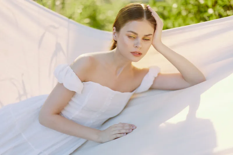 Elegant young woman in a white dress relaxing outdoors, showcasing serenity, nature, and soft lighting for a peaceful atmosphere