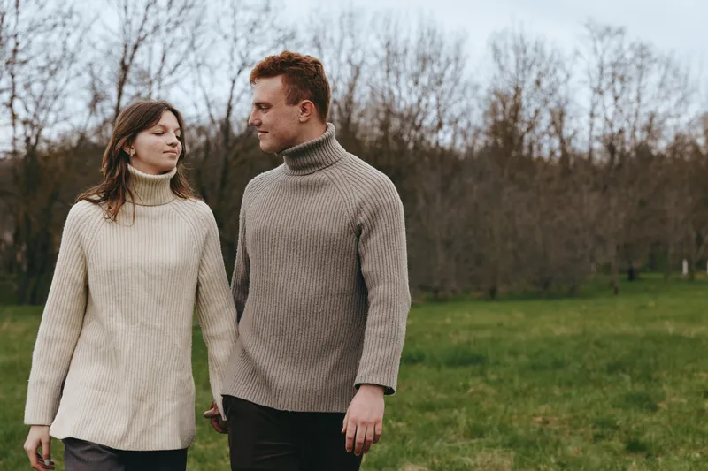 Young couple wearing knitted turtleneck sweaters walk hand in hand through a green field with bare trees in the background, enjoying a calm autumn day outdoors.
