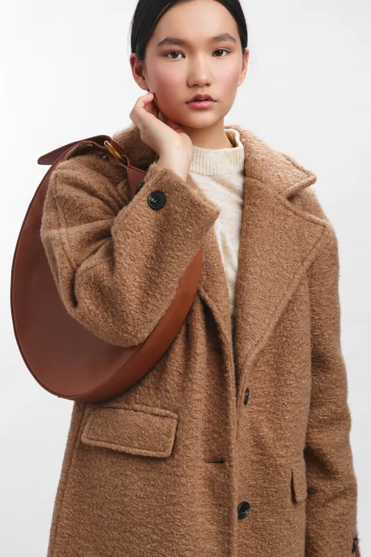 Asian woman wearing a warm brown coat stands in a bright studio, presenting a polished, contemporary fashion look that exudes confidence and poise.