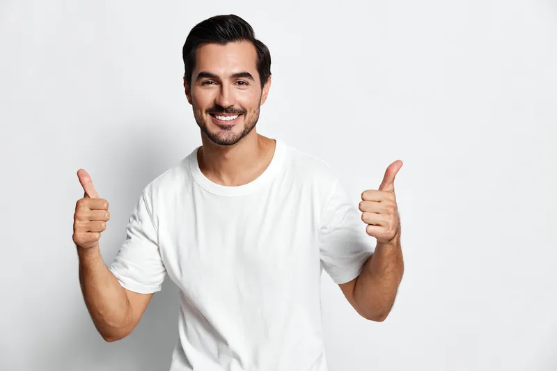 Cheerful smiling young caucasian man in white t-shirt giving thumbs up gesture standing against plain solid light background. Positive emotions and happiness concept.