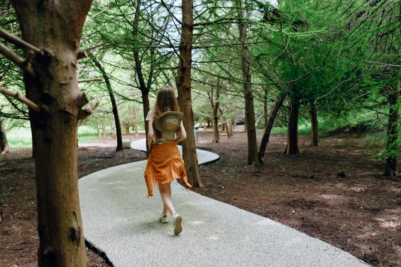 Woman walking forest path with backpack and hat on a gravel trail, nature scene of solo traveler in orange skirt exploring winding walkway among tall trees in peaceful woodland park