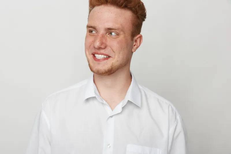 Smiling young man with red hair and beard wearing white shirt looks to side on white background. Casual portrait of happy male model with natural expression and clean studio lighting.