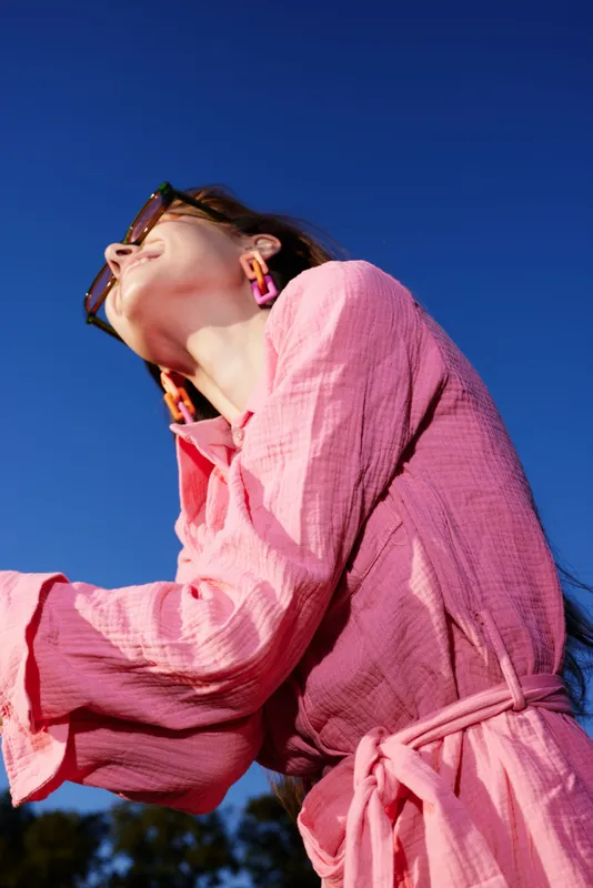 Joyful young woman in a pink dress against a bright blue sky, showcasing a vibrant summer vibe, expressing happiness and freedom with stylish sunglasses