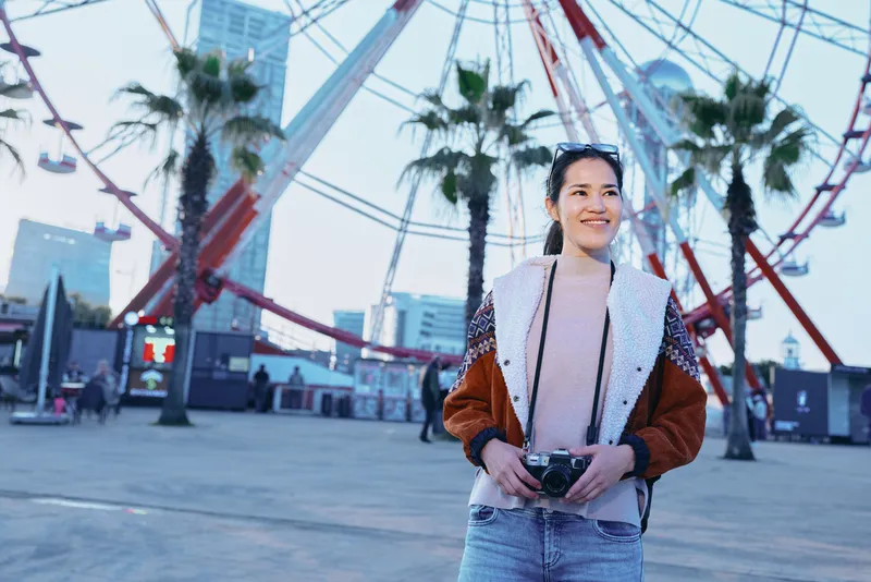A stylish traveler stands in front of a large ferris wheel, holding a camera, wearing a casual jacket and denim, capturing a cheerful outdoor moment at a palm tree lined boardwalk