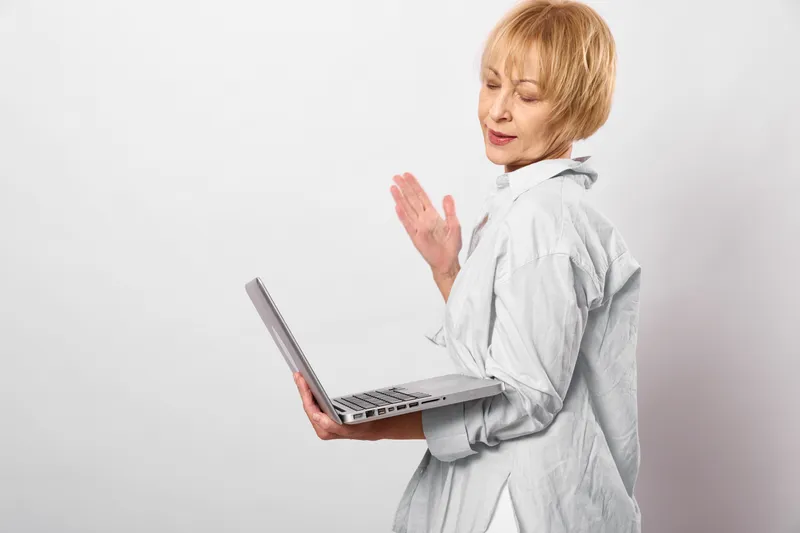 Senior woman with laptop waves hello to camera in bright studio, white background, calm expression, remote work and communication technology concept, copy space