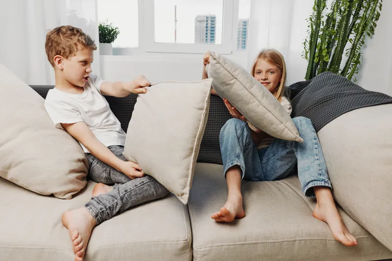 Two children having fun on a beige sofa, playfully hitting each other with pillows in a bright modern living room with casual clothing and natural light.