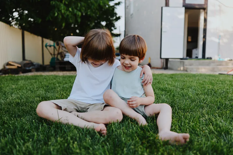 Two cheerful boys sitting on lush green grass, sharing a moment of joy and laughter in a sunny garden, embodying the spirit of childhood and friendship.