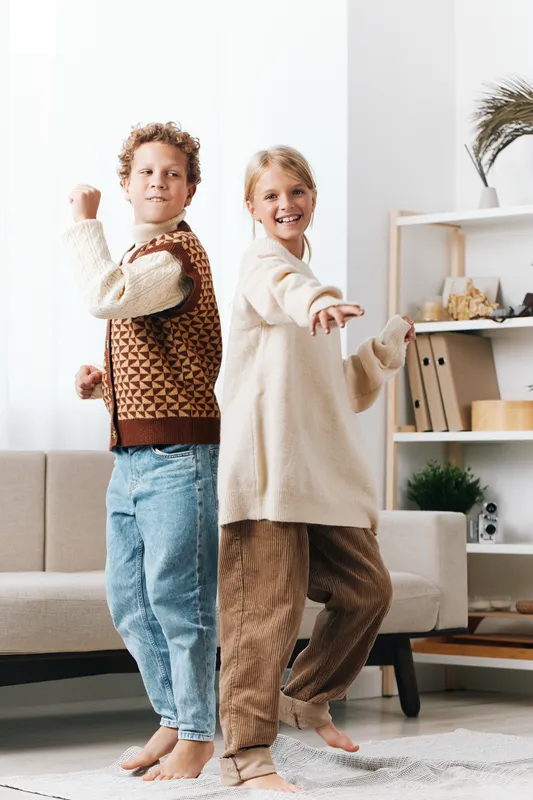 Two children pose back to back in a cozy living room, showcasing playful confidence in patterned sweaters and soft denim, perfect for capturing lighthearted family moments.
