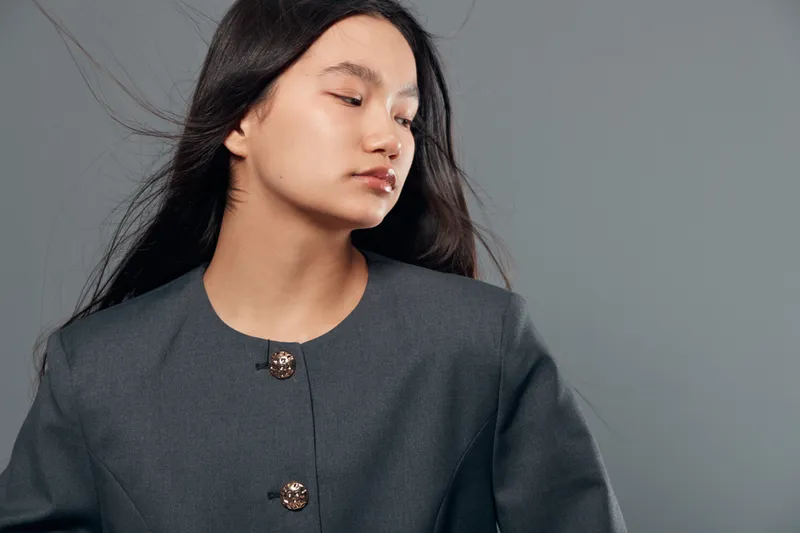 Young woman portrait with long dark hair and closed eyes wearing gray jacket with buttons on gray background. Calm expression, studio shot for fashion or emotion concept with clean style.