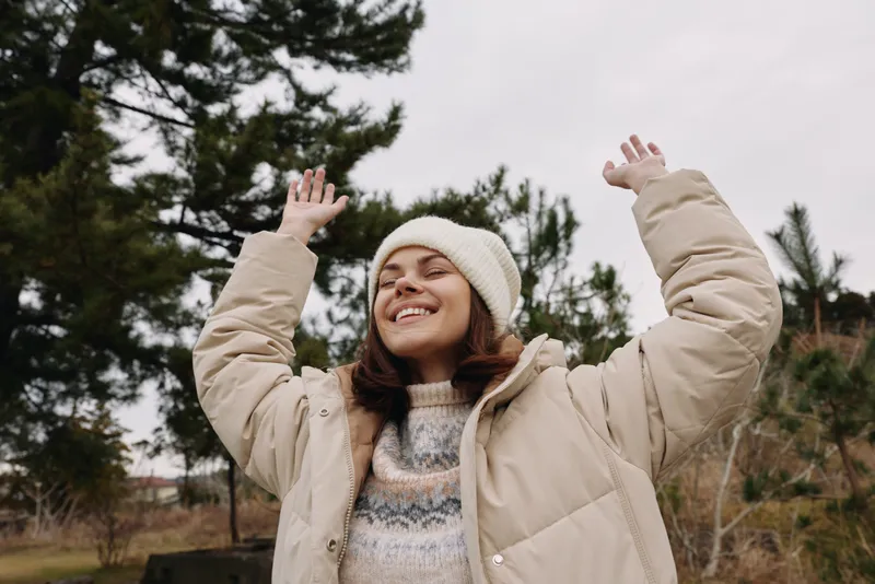 Woman outdoors wearing a warm beige coat and knit sweater, arms raised in joy, smiling under trees on a crisp day, conveying happiness, freedom and winter fashion in a natural park setting