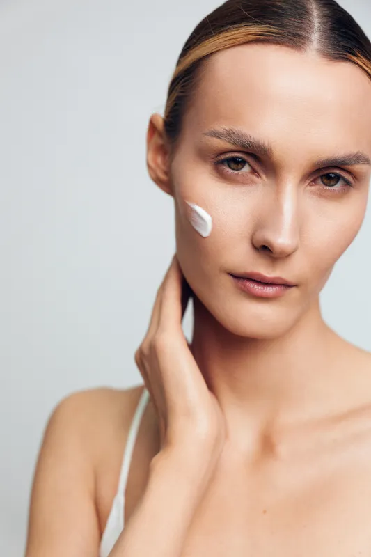 Close up portrait of a woman, smooth face, applying skincare cream, natural makeup, touching her cheek, calm confident gaze, minimal white background, studio light, beauty care