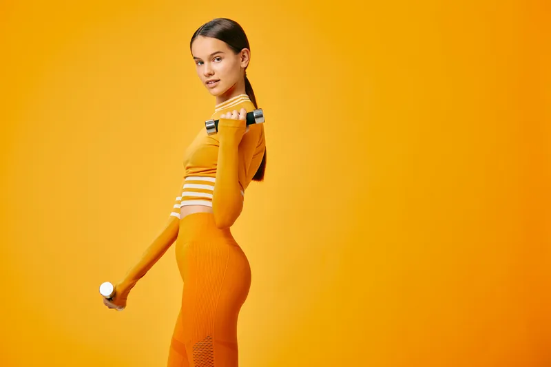 Woman fitness portrait in orange activewear holding dumbbells in studio, sport training pose showing toned muscles, confident posture and dynamic strength against vivid monochrome background.