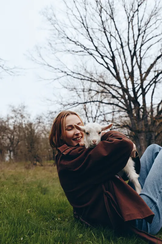 Young woman in a brown jacket happily hugs a white baby goat while sitting on green grass in an autumn park with bare trees in the background.