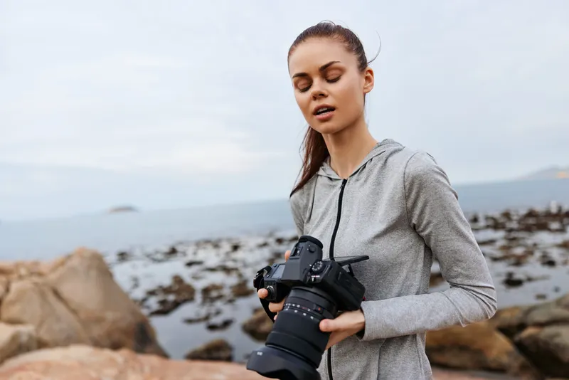 Female photographer in a gray hoodie, capturing scenic views by the sea, showcasing focus and creativity against a serene coastal backdrop