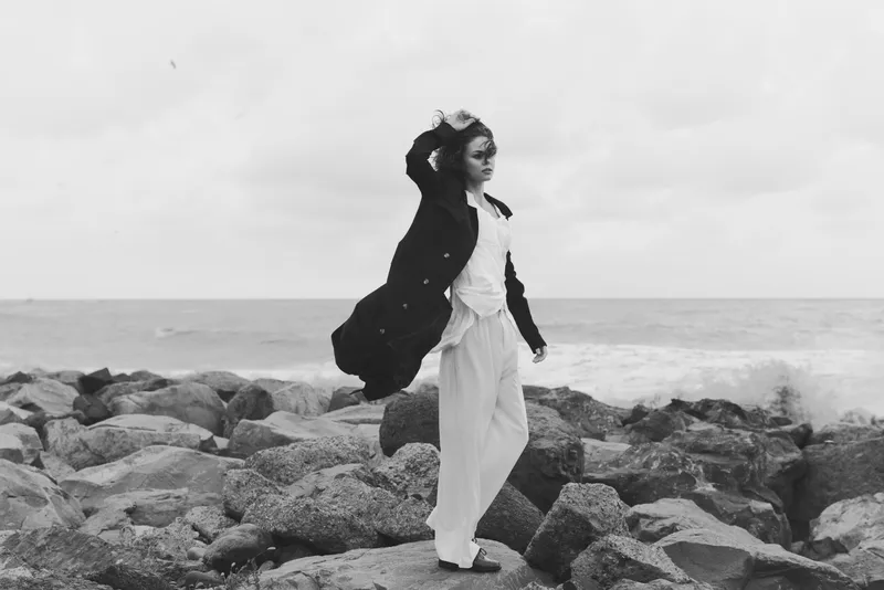 Monochrome scene of a wind swept shoreline with a woman walking on rocks, wearing a long coat and flowing pants, capturing motion, contrast and coastal mood.