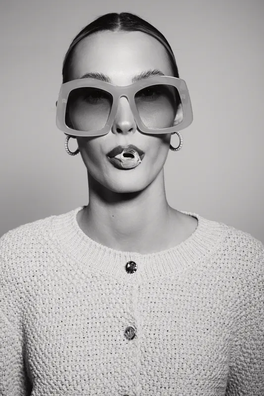 Stylish caucasian woman with fair skin wearing large sunglasses and hoop earrings poses with playful expression in studio. Fashion concept with modern accessories and attitude.