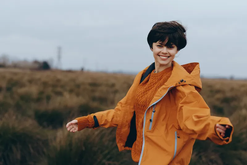 Smiling woman in outdoor jacket and scarf walking in a nature field, autumn landscape under cloudy sky, casual lifestyle portrait, warm orange clothing, soft daylight, open countryside.