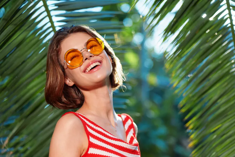 A joyful young woman wearing round orange sunglasses and a red striped top stands among lush palm leaves. Bright sunshine highlights a carefree, summery mood and relaxed, optimistic energy.