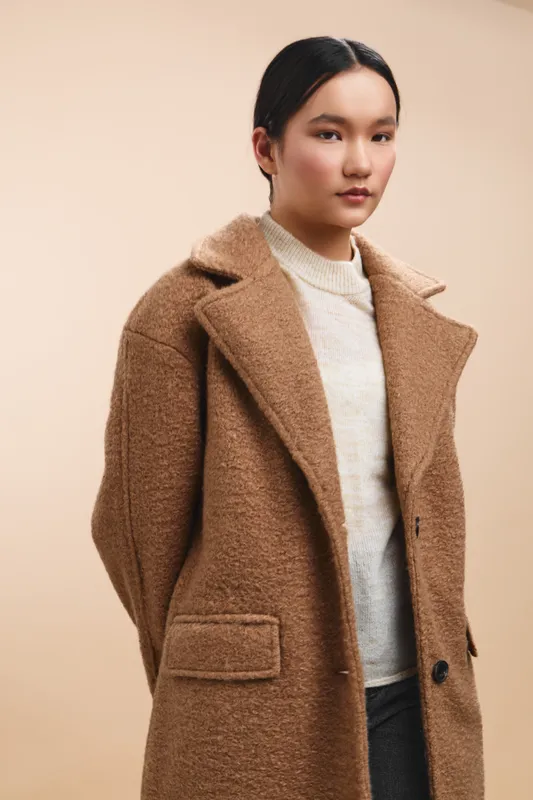 asian woman wearing a tan woolen coat stands against a soft beige backdrop, presenting a stylish, contemporary look that conveys confidence and warmth.