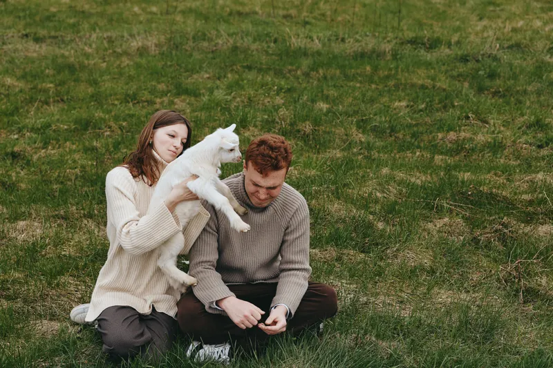 Couple outdoors with a playful goat kid on a grassy field, capturing a warm moment of affection and connection. Natural light and relaxed mood emphasize lifestyle, care, and rural charm in this