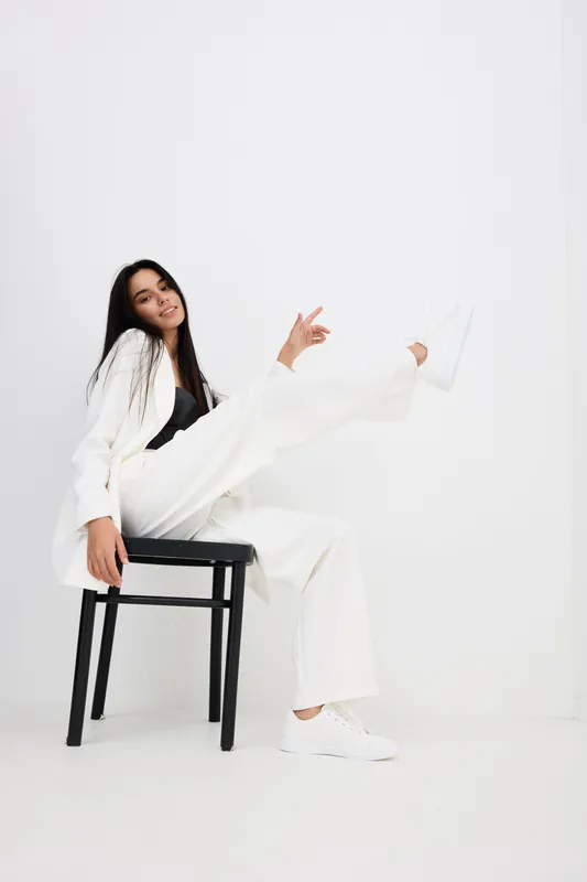 Young woman sitting on chair with one leg raised, wearing white suit and sneakers, displaying relaxed and playful mood in minimalist studio setting.