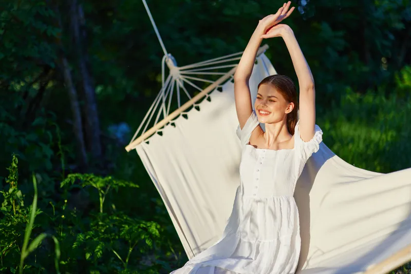 A young woman in a white dress stretching joyfully in a hammock, surrounded by greenery, evoking a sense of relaxation and tranquility in a natural setting