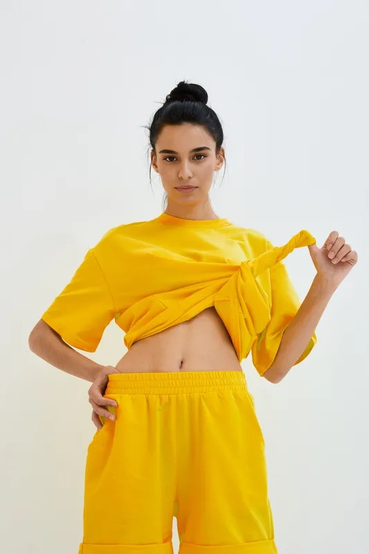 young woman in yellow casual outfit lifting shirt revealing stomach, standing against white background with confident expression and bun hairstyle