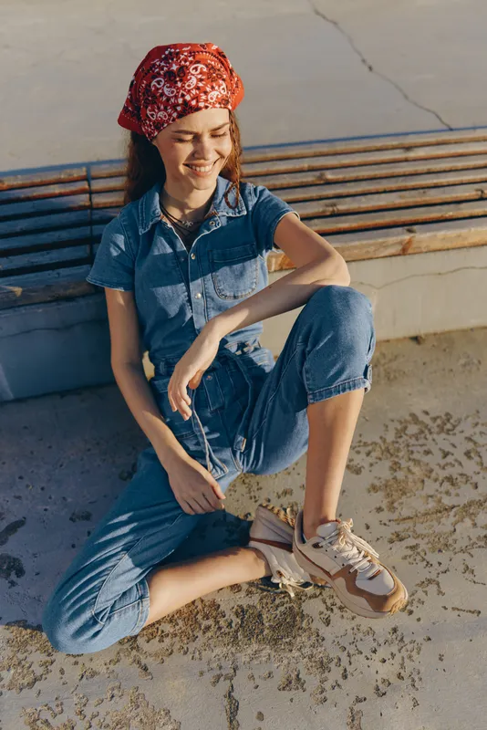 Lifestyle woman in modern boho-western denim street style with red bandana and casual sneakers, relaxed outdoors with warm film color tones and natural sunlight.