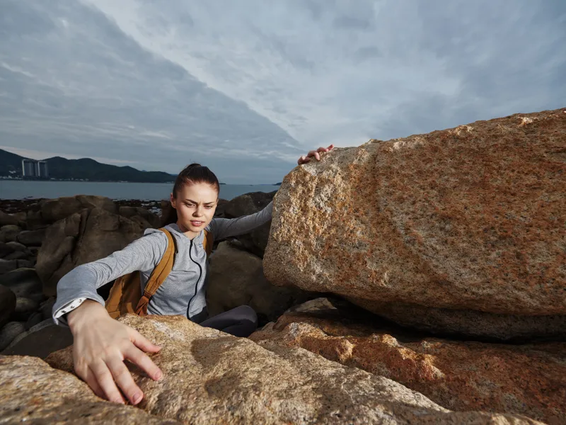 Young woman climbing rocky coastline, wearing gray athletic wear, expressing determination, against a dramatic sky and ocean background, ideal for adventure themes