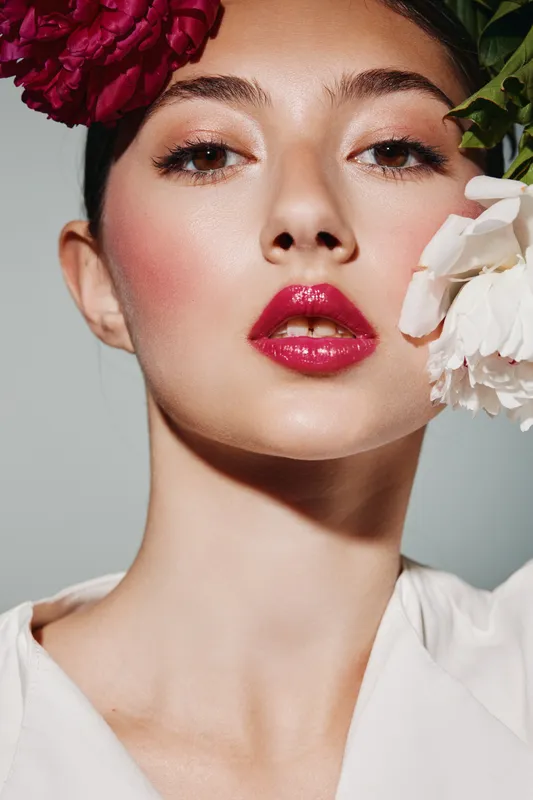 Close-up of a young woman with vibrant red flowers around her face and striking bright pink glossy lips. Soft lighting and clean grey background enhance the fashion beauty look.