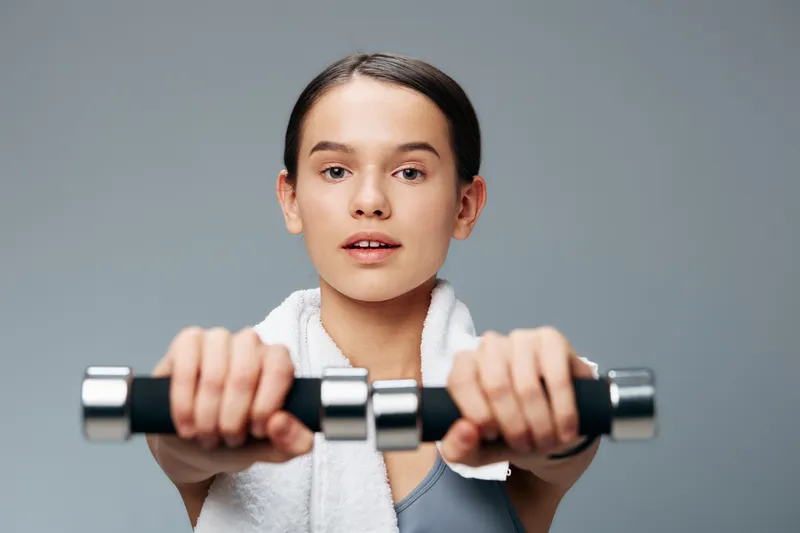 Fitness woman in a gym holds dumbbells, arms extended, focused expression, towel around her neck, intense training and exercise in a bright studio.