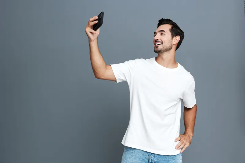 Happy smiling young man holding smartphone and taking selfie in casual white t-shirt against plain grey background. Modern lifestyle concept.