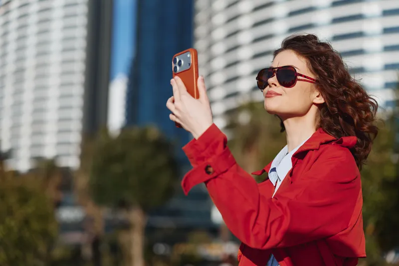 Cheerful woman outdoors taking a selfie in a red coat, sunglasses, urban backdrop, sunlit moment, candid portrait, modern city vibe, bright