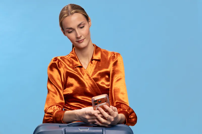 Young woman in shiny orange blouse uses smartphone sitting on suitcase against blue background. Concept of travel, communication, and modern lifestyle with casual fashion and technology.