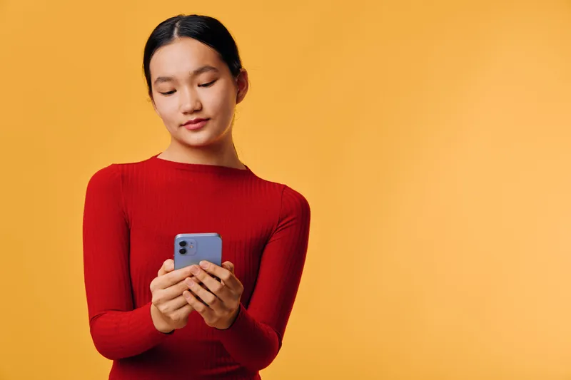 Young woman in red sweater using smartphone for texting or browsing on yellow background. Female holding device with focused expression for technology and communication concept.