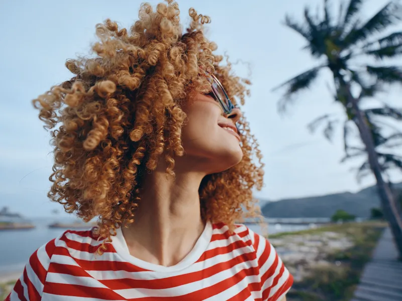 Closeup portrait of a joyful person with thick curly hair, sunglasses, and a striped shirt, looking upward with a bright mood against a beachside backdrop.