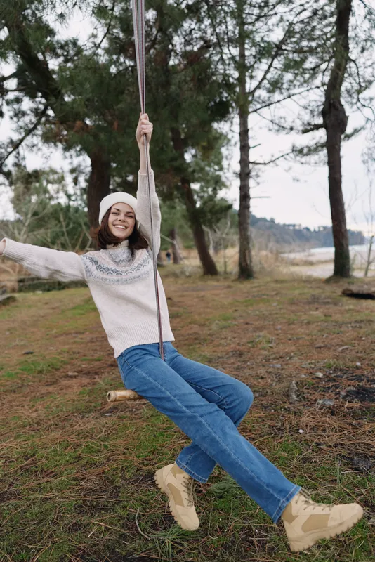 Woman on a rope swing outdoors among trees, wearing a knitted sweater, jeans and boots, enjoying a carefree moment in nature.
