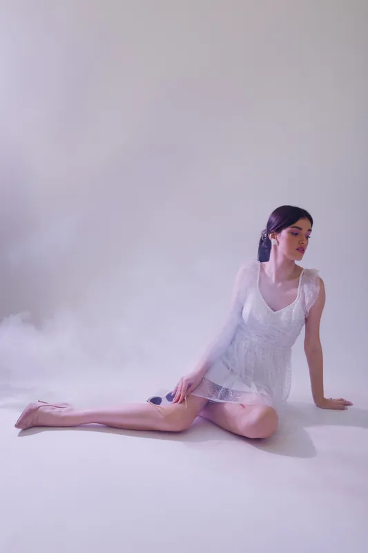 Young woman in a white dress sits on the floor with one leg extended and looks to the side. Soft lighting and mist create a dreamy atmosphere. Concept of beauty fashion and elegance.