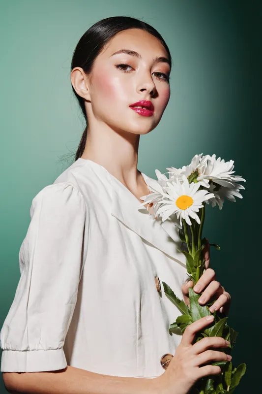 Young woman with glossy red lips holds white daisies against a light green background. Soft fashion lighting highlights her smooth skin and modern, elegant style posing beauty concept.
