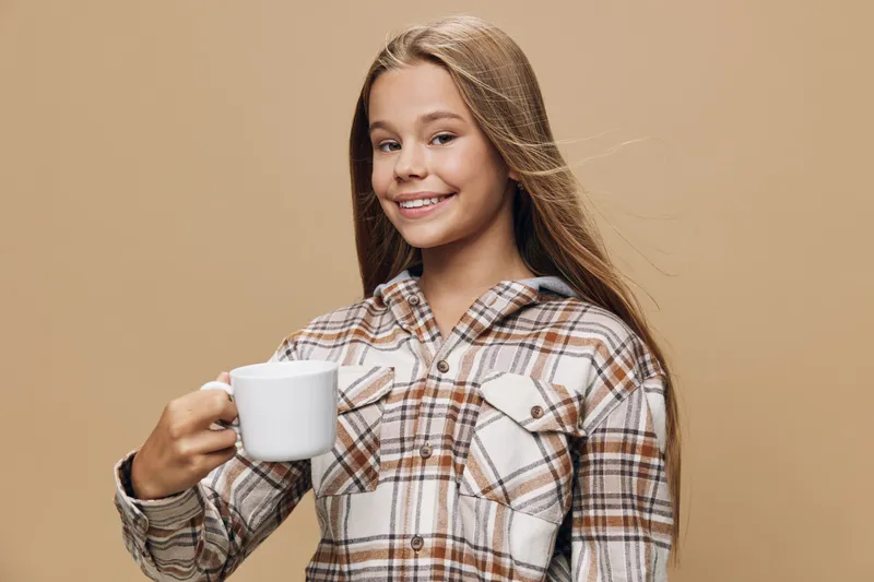 Girl smiling and holding white ceramic cup wearing checked shirt on beige background, lifestyle portrait of happy female drinking beverage with relaxed expression and long hair.