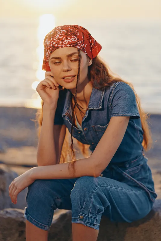lifestyle woman in modern boho-western denim street style wearing red bandana, relaxed outdoors by water with warm film color lighting, casual summer mood