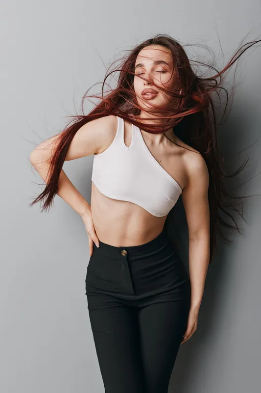 Young woman posing with closed eyes and flowing hair wearing white crop top and black pants against gray background. Fashion, beauty, casual style, confident, studio portrait with movement.